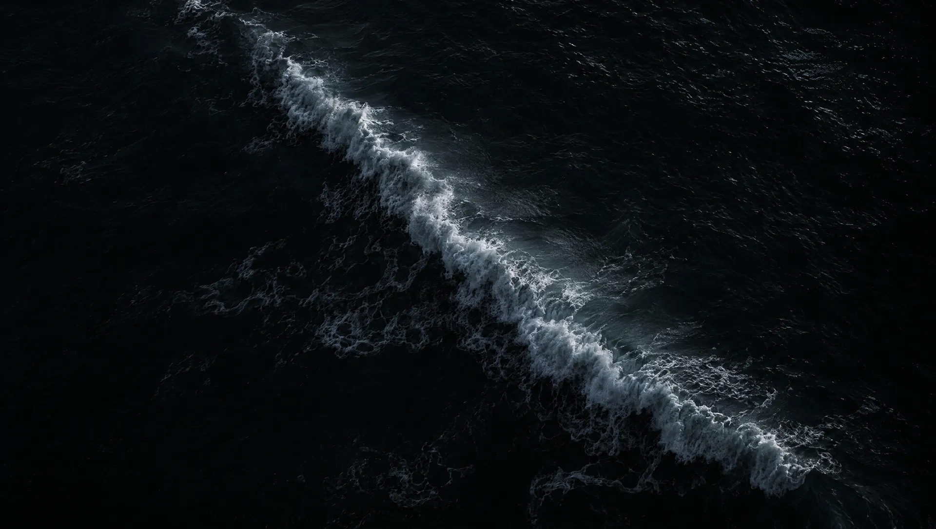Open ocean from above at night, one wave breaking across black water.
